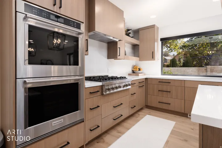 Stainless steel double oven, gas range, wood-finish cabinetry with black hardware, white countertops, and a large window