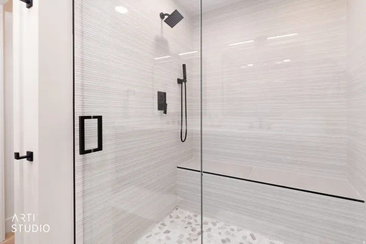 Walk-in shower with clear glass enclosure, matte black fixtures, stone pebble flooring, integrated bench, and textured wall tiling
