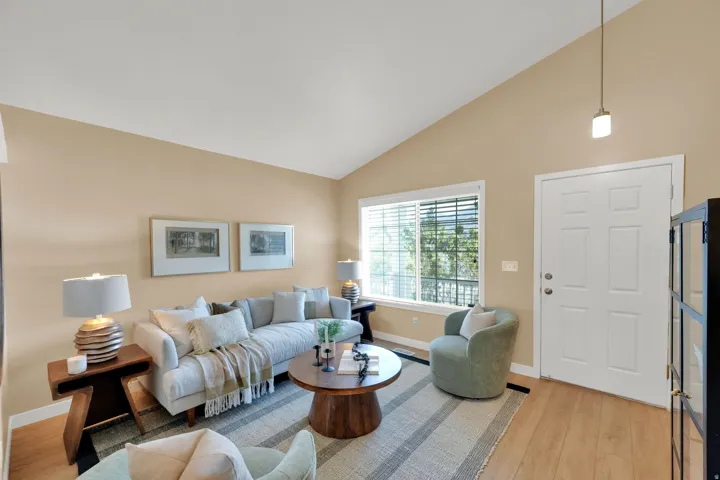 Vaulted ceiling with an accent pendant light, expansive window with white blinds, light wood-finish flooring, and a paneled entry door