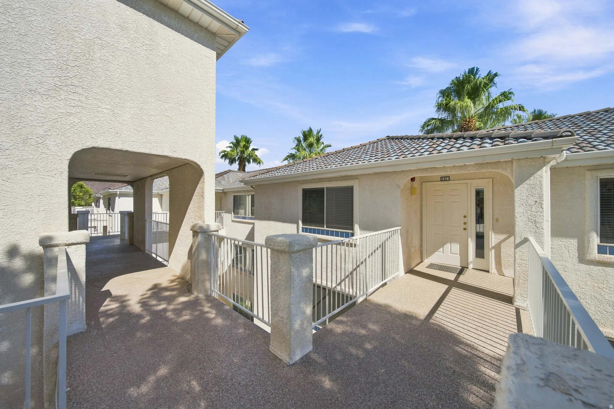 Stucco exterior with a tiled roof, featuring a covered entryway, a white door with a clear glass side panel, and metal railings