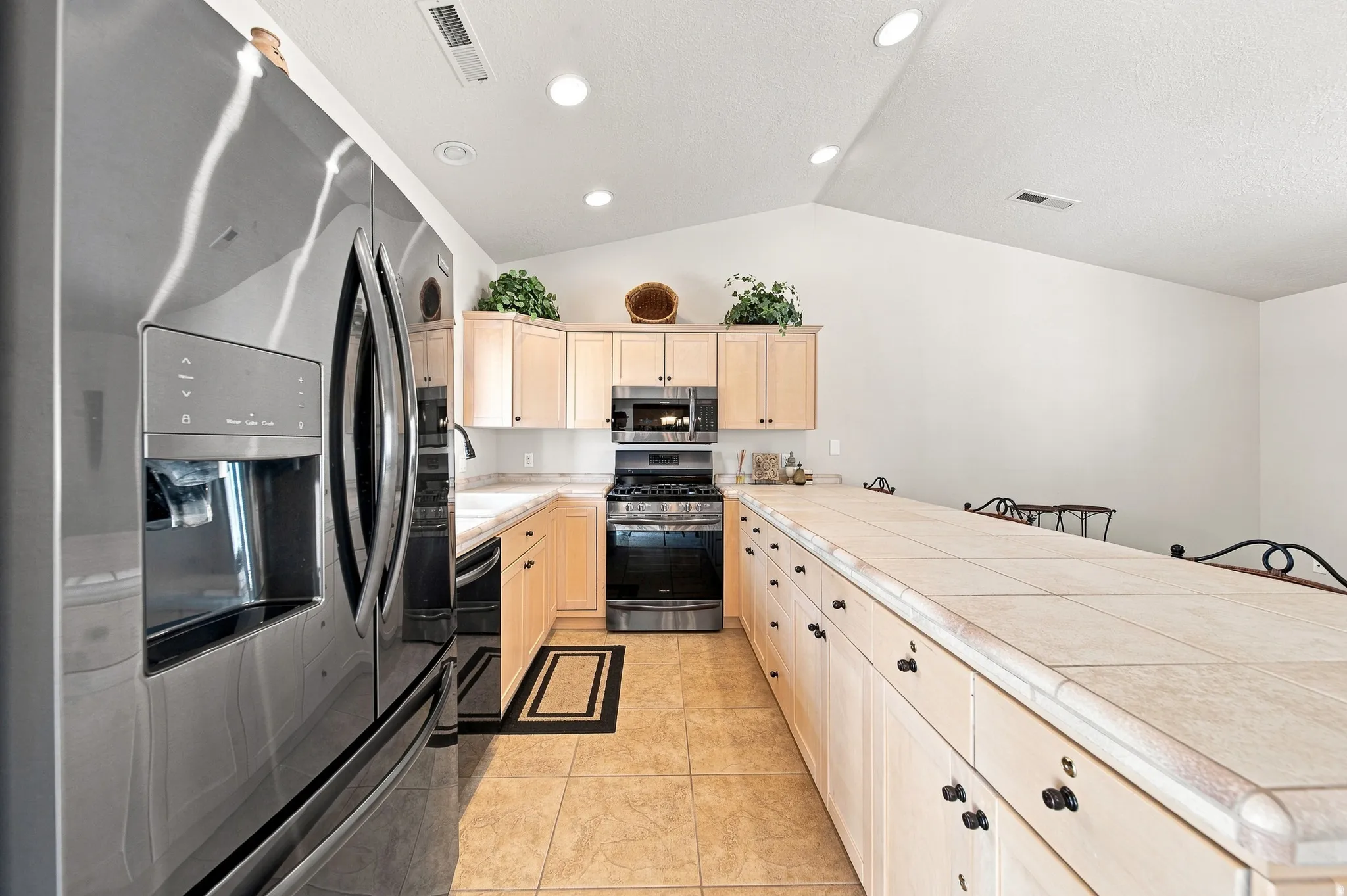 Stainless steel French door refrigerator, light wood-finish cabinetry, tile flooring, tile countertops, and vaulted ceilings