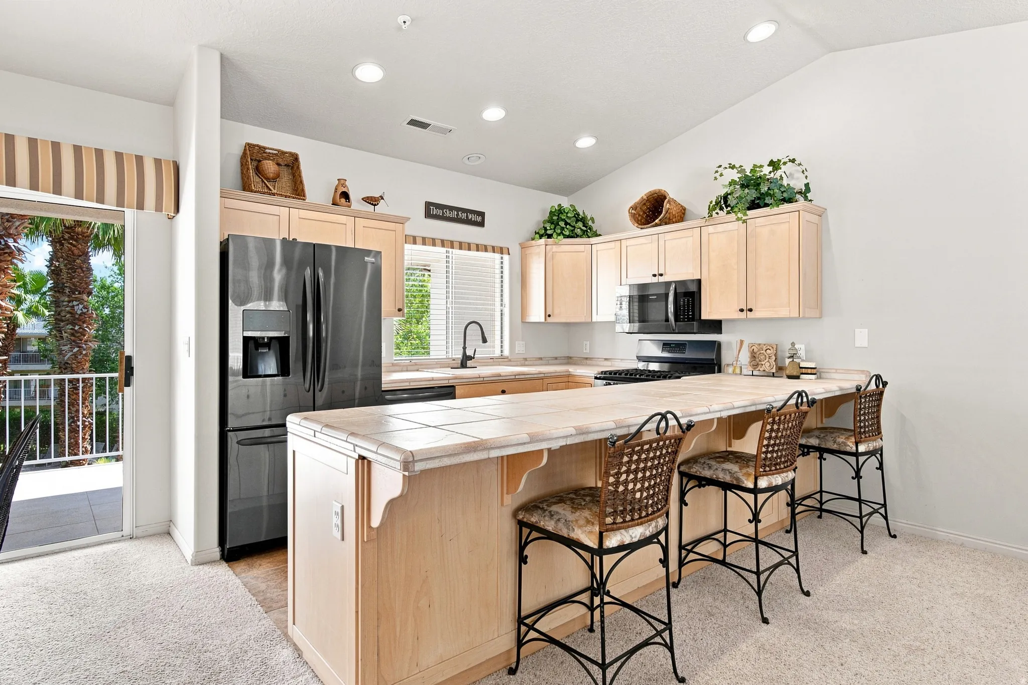 Kitchen featuring recessed lighting, a vaulted ceiling, a tile-top island with seating, light wood cabinetry, and a sliding glass door