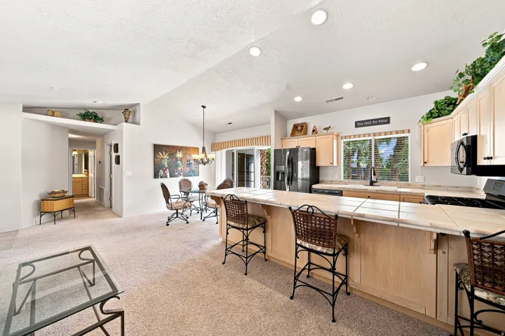 Vaulted ceilings and recessed lighting illuminate the open-concept living space, featuring a kitchen with light wood-finish cabinetry, a large breakfast bar with tiled countertop, and a dining area with a chandelier