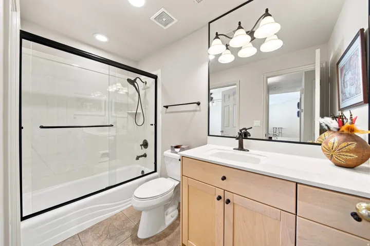 Bathroom featuring a full vanity with an integrated sink and light wood cabinetry, a large framed mirror, a five-light fixture, and a shower-over-tub with a sliding glass enclosure and black hardware