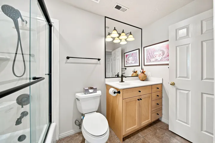 Glass-enclosed shower with dark bronze fixtures, light wood-finish vanity with white countertop, dark bronze framed mirror, and tiled flooring