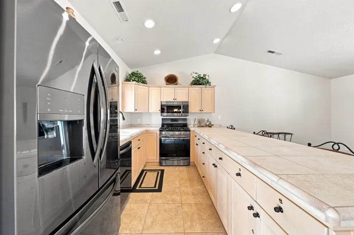 Stainless steel French door refrigerator, light wood-finish cabinetry, tile flooring, tile countertops, and vaulted ceilings