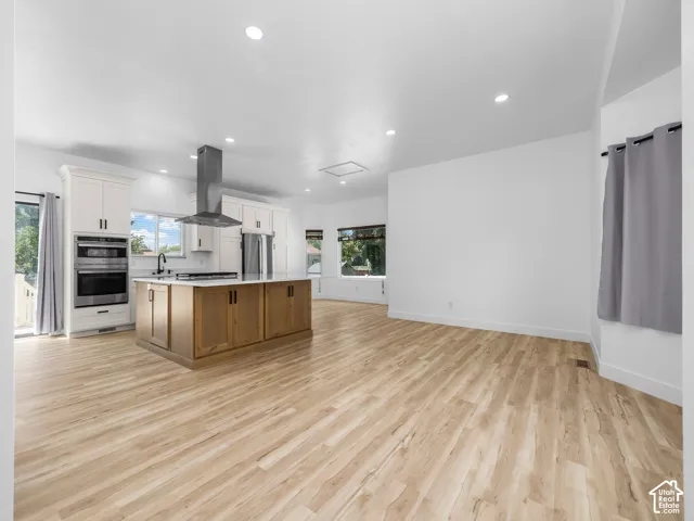 Open-concept kitchen and dining area featuring wood-finish flooring, a substantial wood-grain island with seating, stainless steel appliances including a wall-mounted range hood, recessed ceiling lighting, and abundant natural light from multiple windows