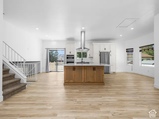 Open-concept kitchen and living area featuring wide-plank wood-finish flooring throughout, a substantial island with wood cabinetry and stainless steel appliances, white painted cabinetry with integrated storage, and a stainless steel range hood