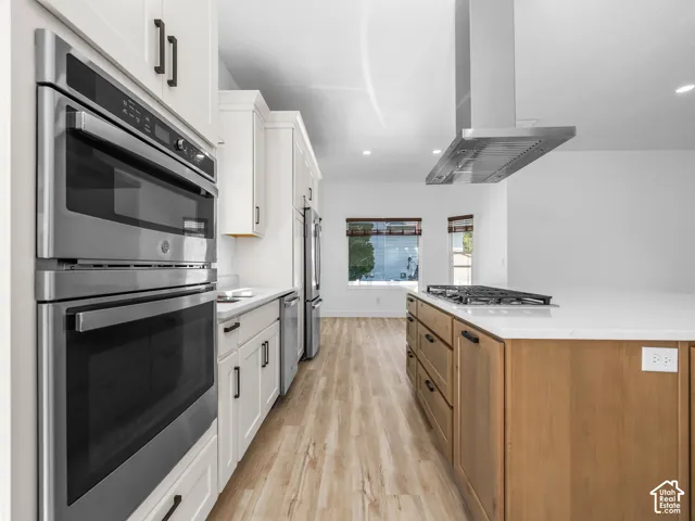 Modern kitchen featuring stainless steel double wall ovens and matching refrigerator, white cabinetry with hardware accents, wood-finish island with prep space, white quartz countertops, gas cooktop with stainless steel range hood, and warm wood-finish fl