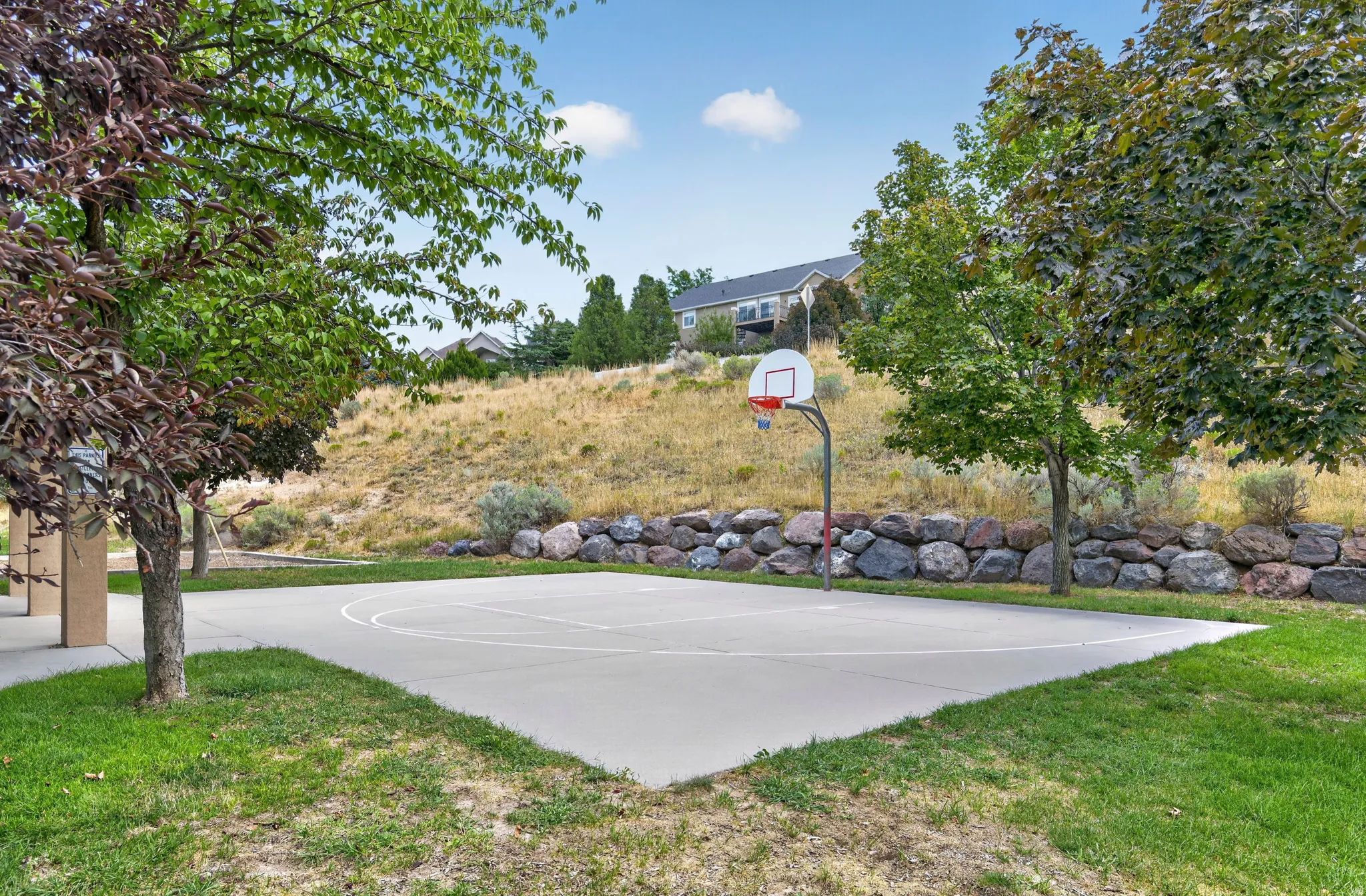 Paved basketball court with a hoop, surrounded by green grass and mature trees, featuring a natural rock retaining wall