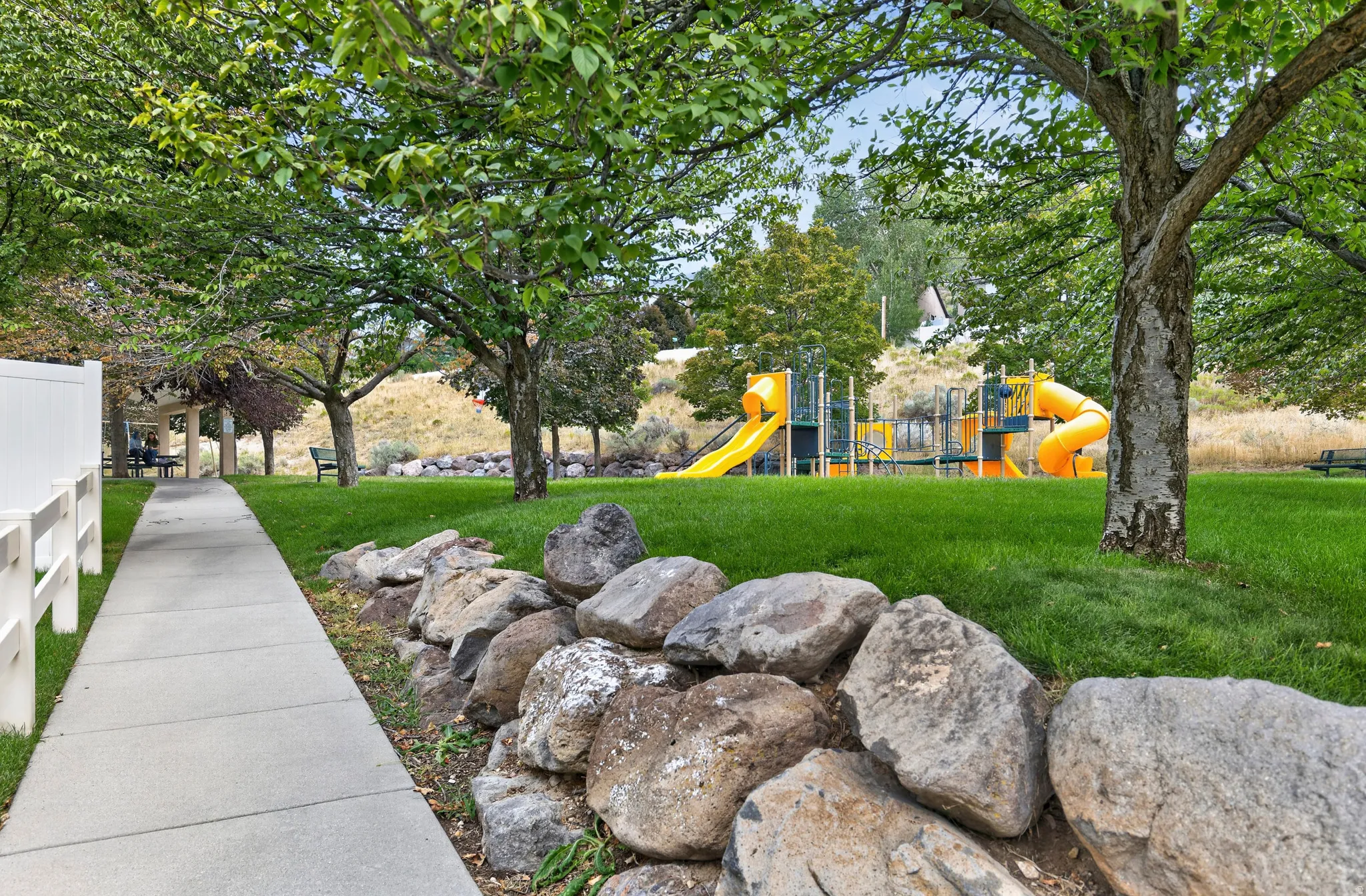 Community playground with multiple slides, rock retaining wall, mature trees, and a paved walkway