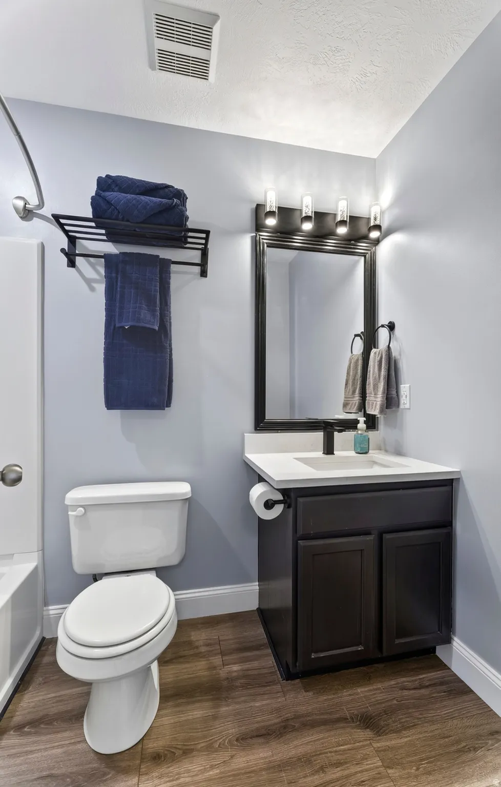 Wood-finish flooring, dark wood vanity with white countertop, black-framed mirror, and a modern light fixture