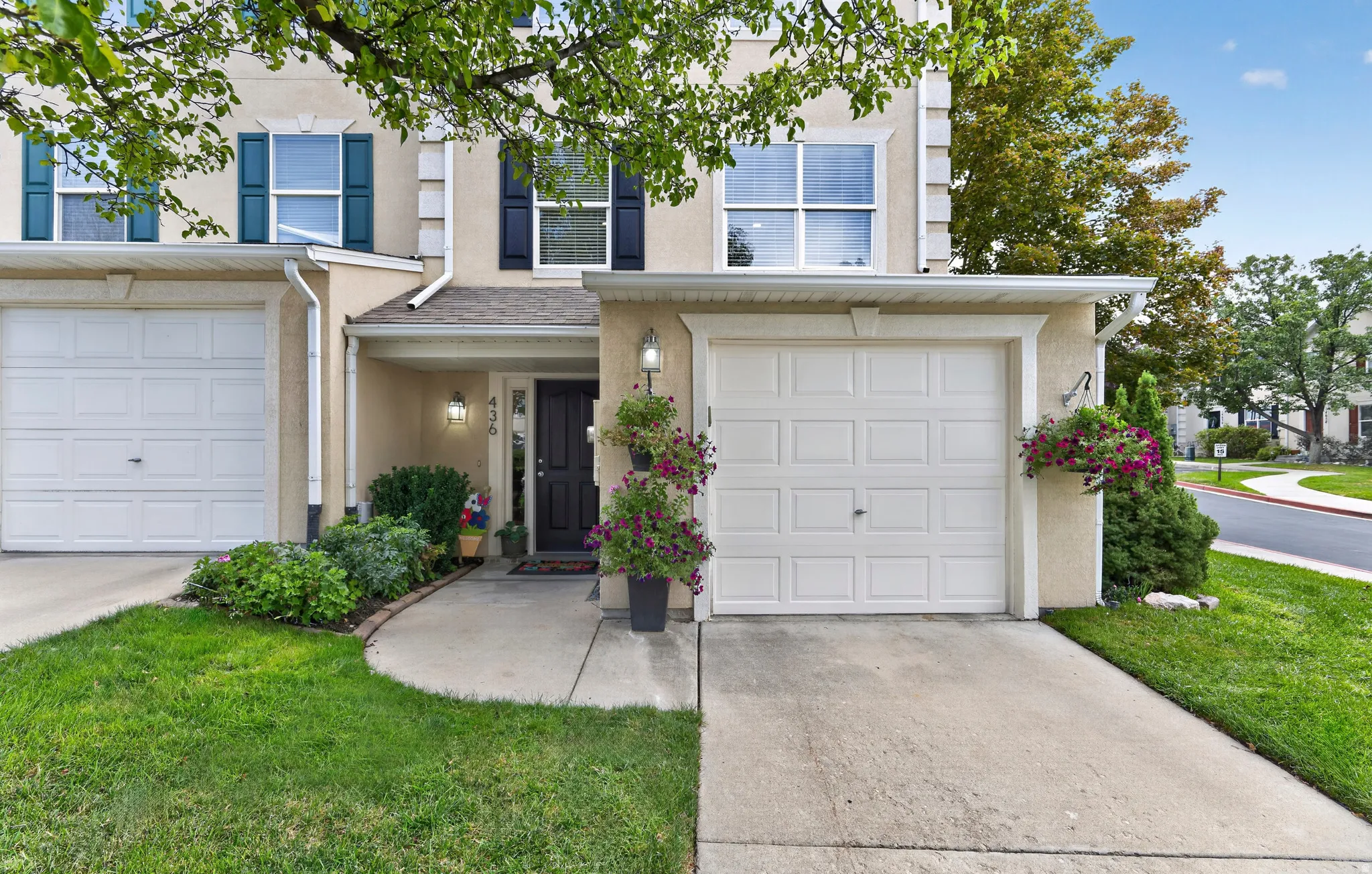 Two-car garage home with white panel doors, neutral exterior featuring stone and siding accents, manicured front lawn with mature landscaping, welcoming entry with planters and sconce lighting, and tree-lined streetscape setting