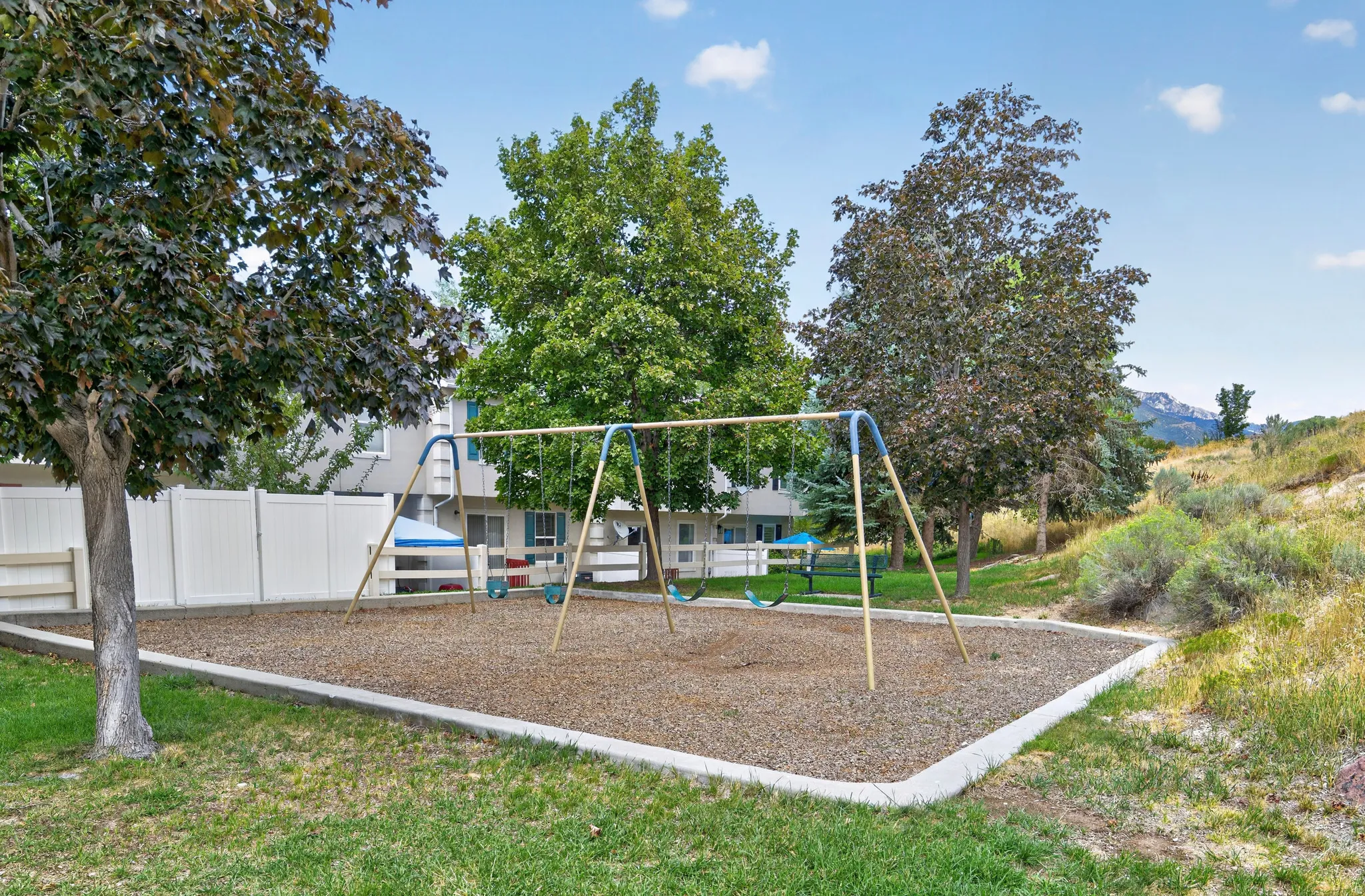 Community playground featuring a swing set on a wood chip surface, surrounded by mature trees and manicured grass