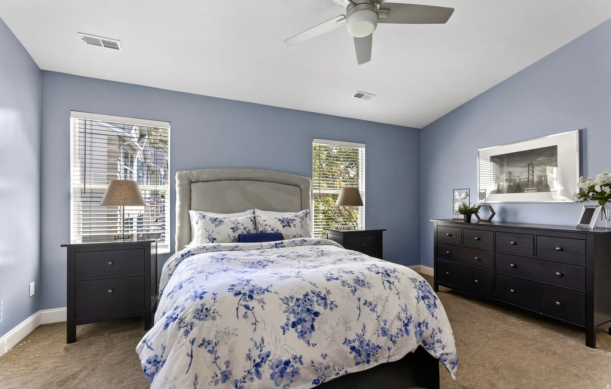 Serene primary bedroom showcasing soft blue-gray walls, a upholstered headboard with nailhead trim, black wood nightstands and dresser, ceiling fan, and abundant natural light from dual windows with plantation shutters