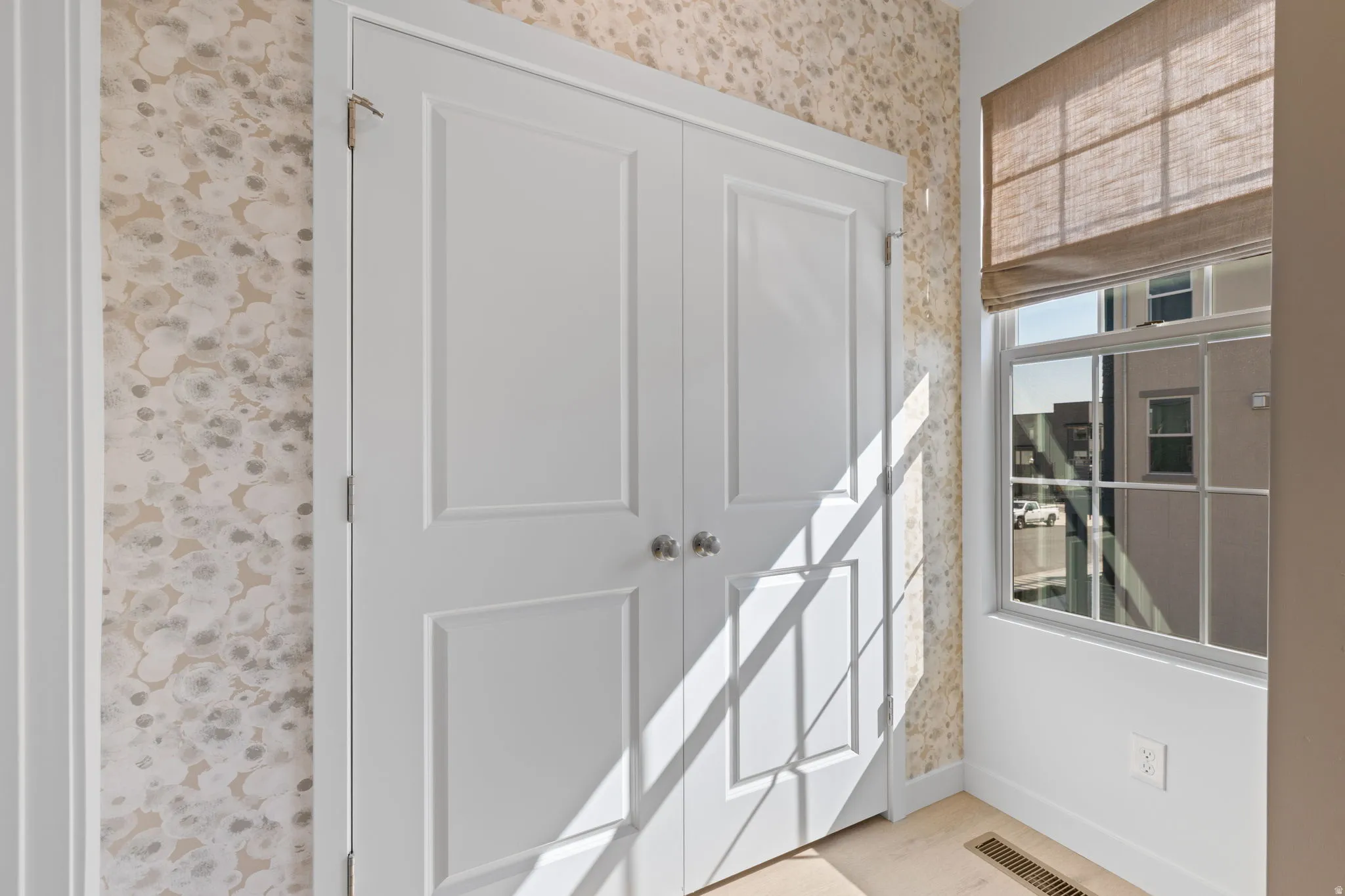 Double panel doors, patterned wallcovering, large window, woven Roman shade, and light wood-finish flooring