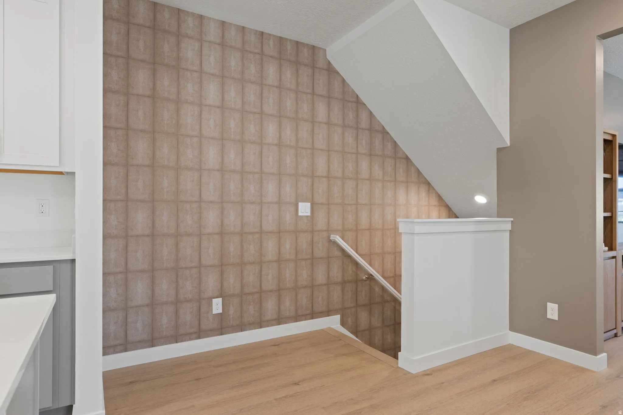 Wood-finish flooring, staircase with white railing, patterned accent wall, white wall cabinetry, and light countertop