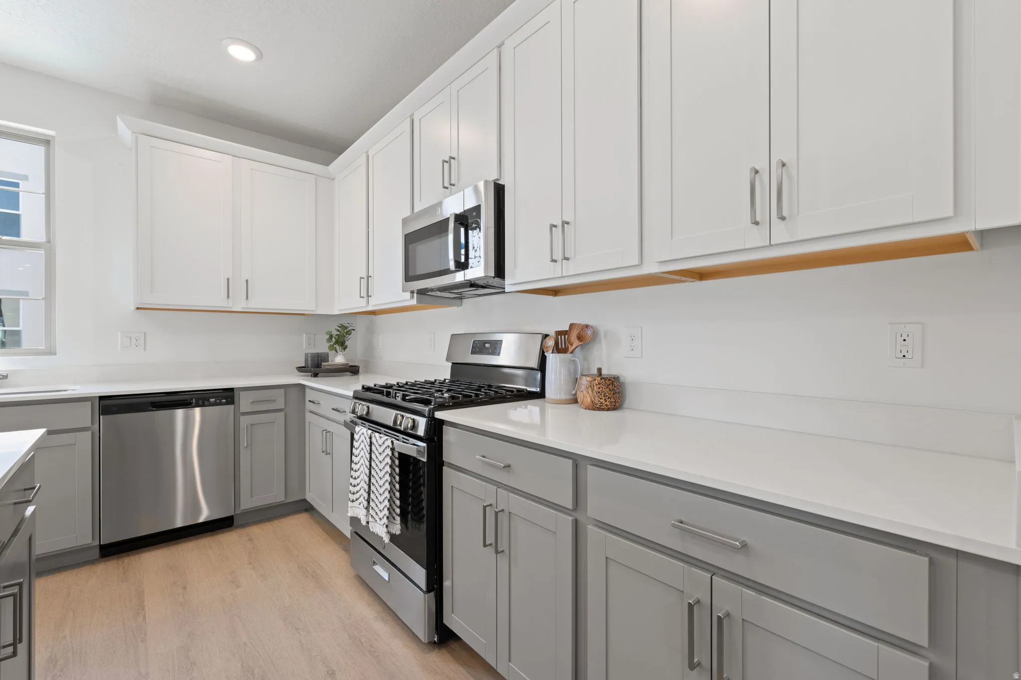 Modern kitchen featuring white shaker-style cabinetry with brushed nickel hardware, stainless steel appliances including a gas range and dishwasher, quartz countertops, a wood-finish accent rail, and recessed lighting throughout
