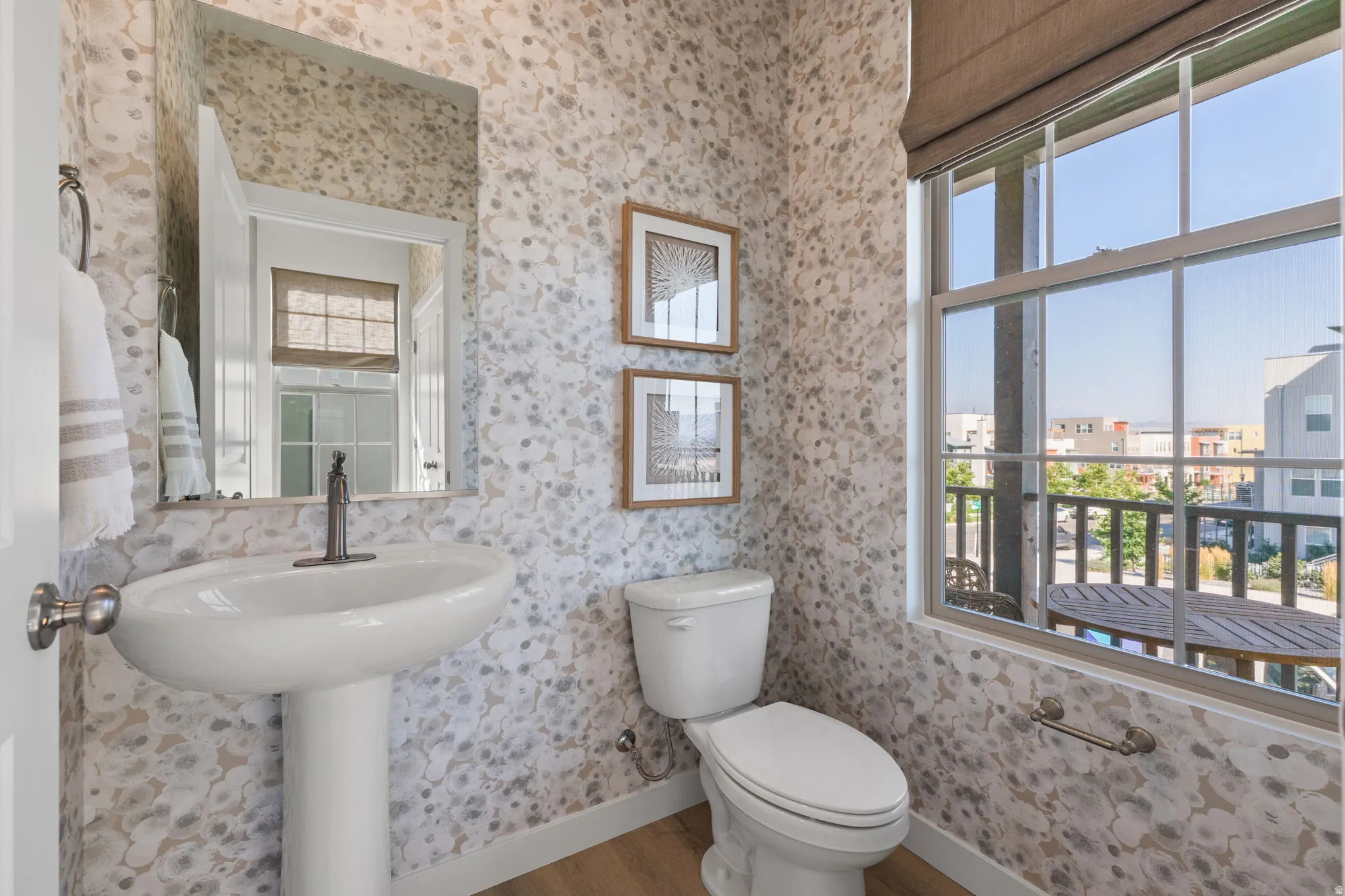 Pedestal sink with a dark bronze-finish faucet, framed mirror, decorative wall covering, and a window with a roller shade