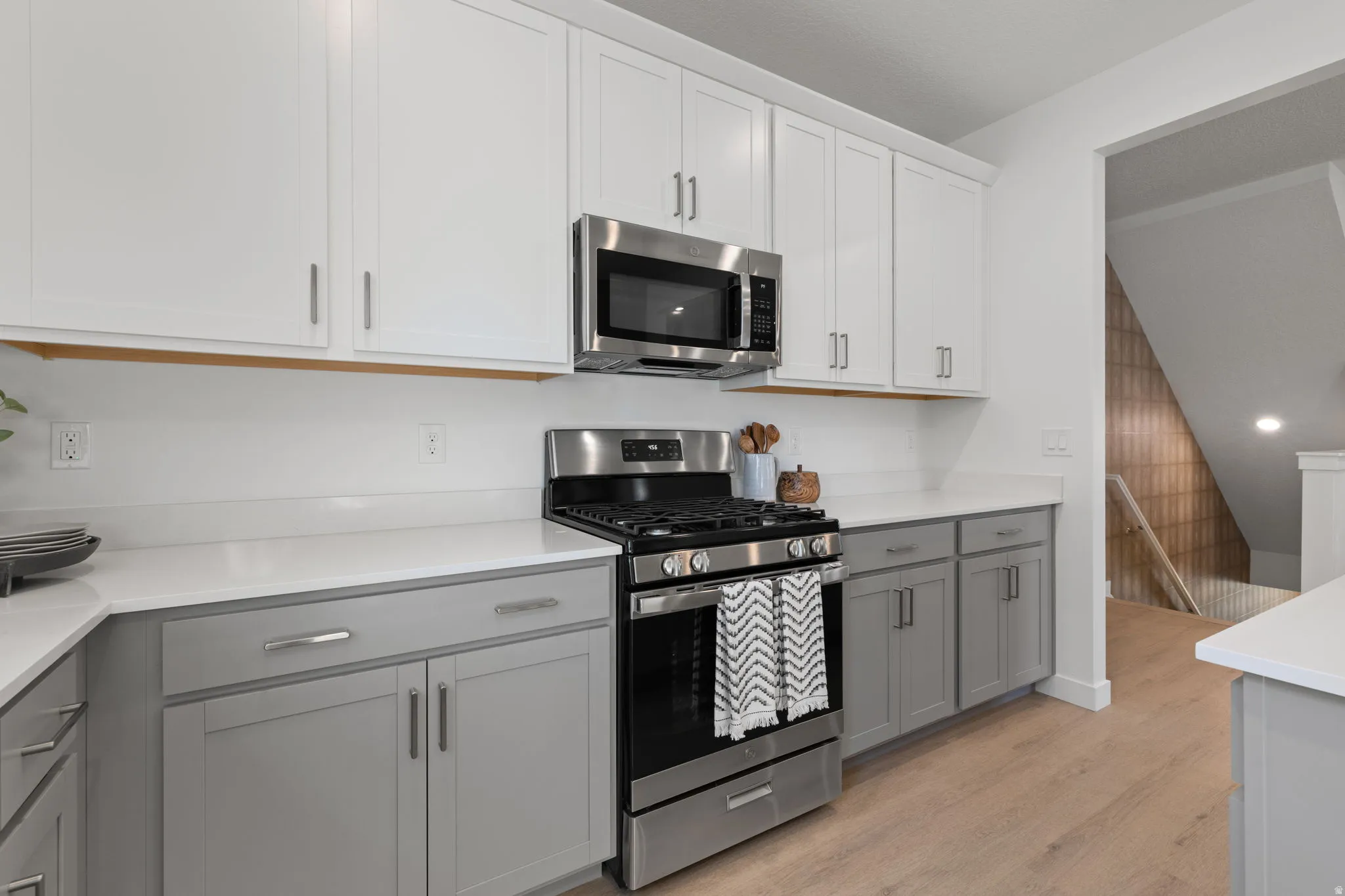 Two-tone cabinetry with white upper and gray lower cabinets, stainless steel appliances, white countertops, wood-finish flooring, and a built-in microwave