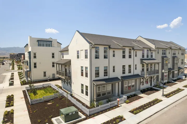 Modern townhome exterior featuring light-colored siding, dark-toned roofing, multiple balconies, a front patio, and a fenced yard with a manicured lawn