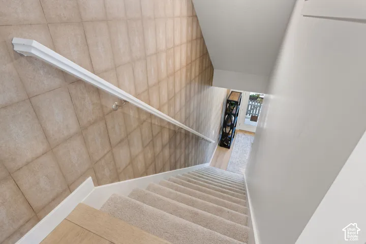 Stairwell featuring carpeted treads, wood-finish risers, and a white handrail set against a tiled wall