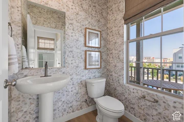 Pedestal sink with a dark bronze-finish faucet, framed mirror, decorative wall covering, and a window with a roller shade