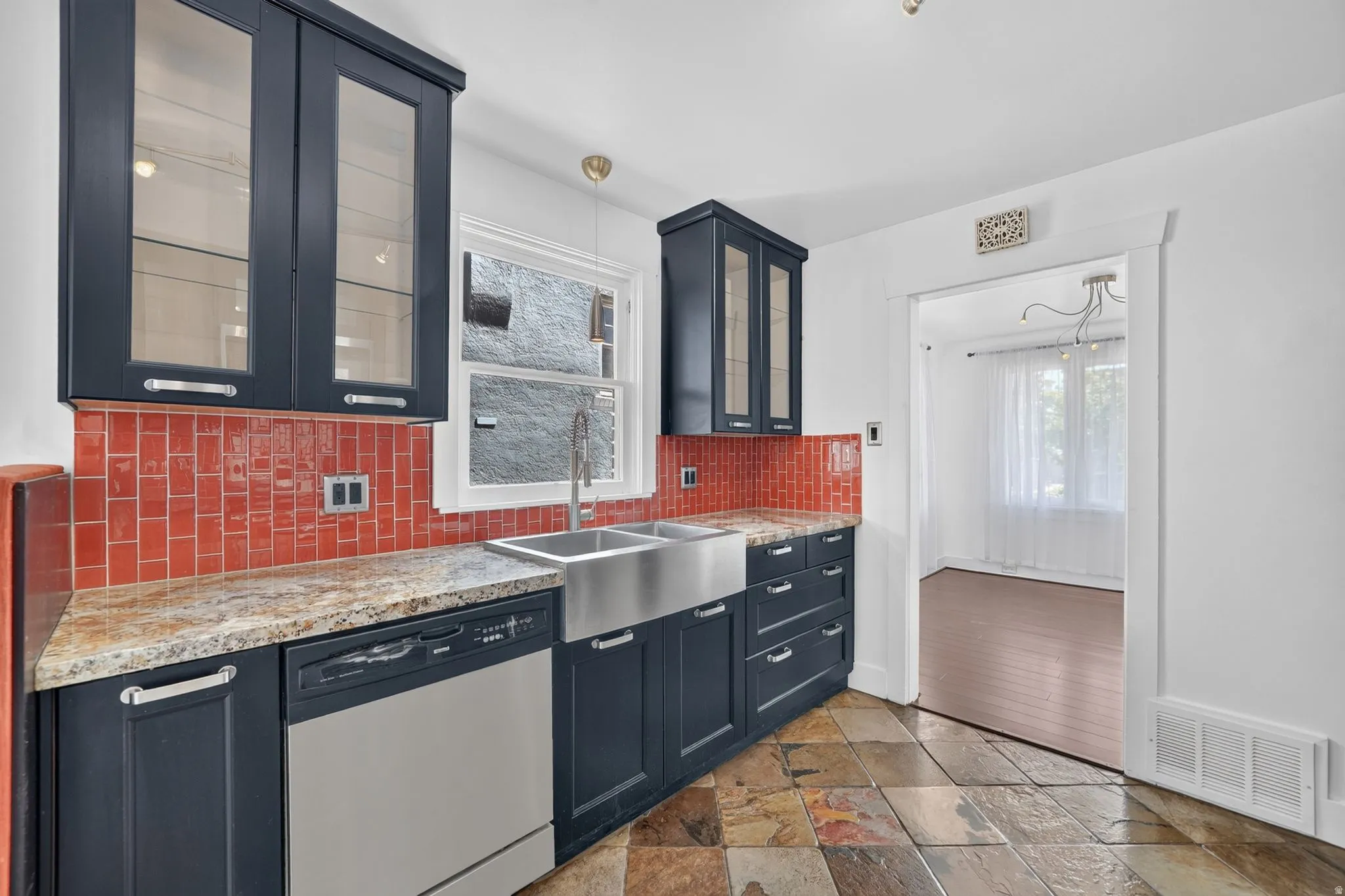 Kitchen featuring slate tile flooring, dark cabinetry with glass-front uppers, granite-style countertops, a stainless steel apron sink, and a red subway tile backsplash
