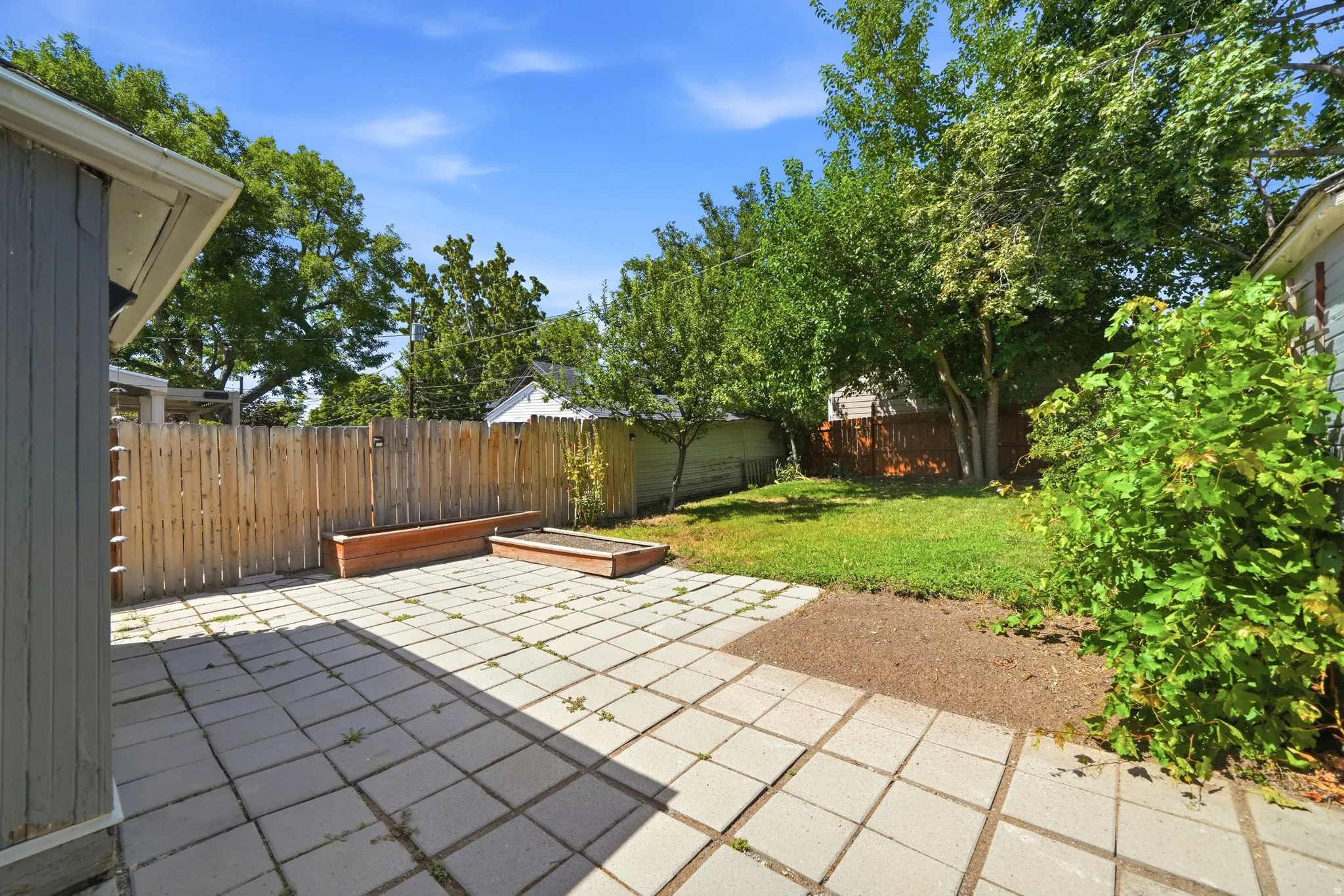 Expansive patio area with large square pavers, featuring a wooden privacy fence, raised garden beds, and a grass lawn with mature trees