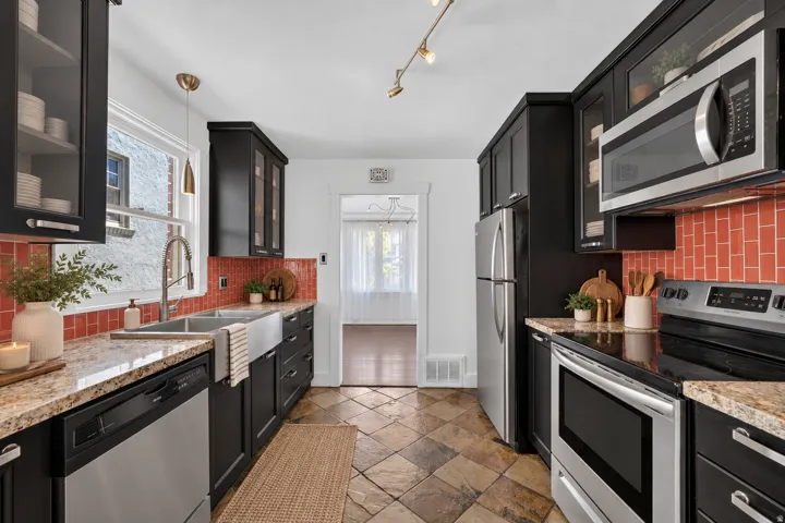 Kitchen featuring black cabinetry, granite countertops, stainless steel appliances, a farmhouse sink, and a red subway tile backsplash