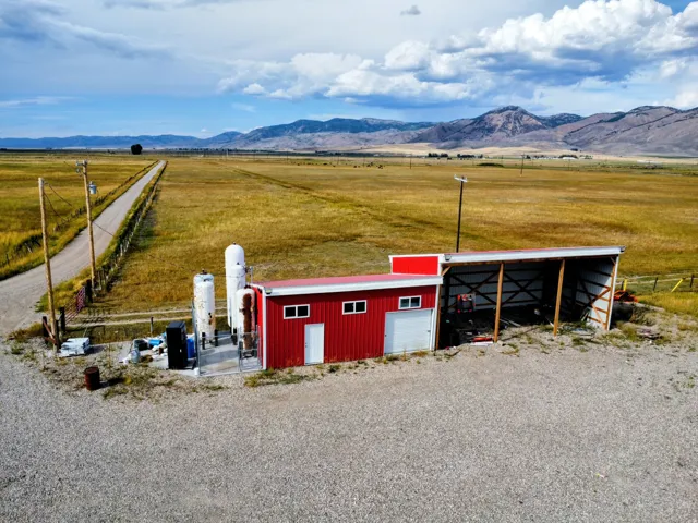 Red-sided utility building with white trim, an attached open-sided lean-to structure, a gravel lot, and a long gravel driveway
