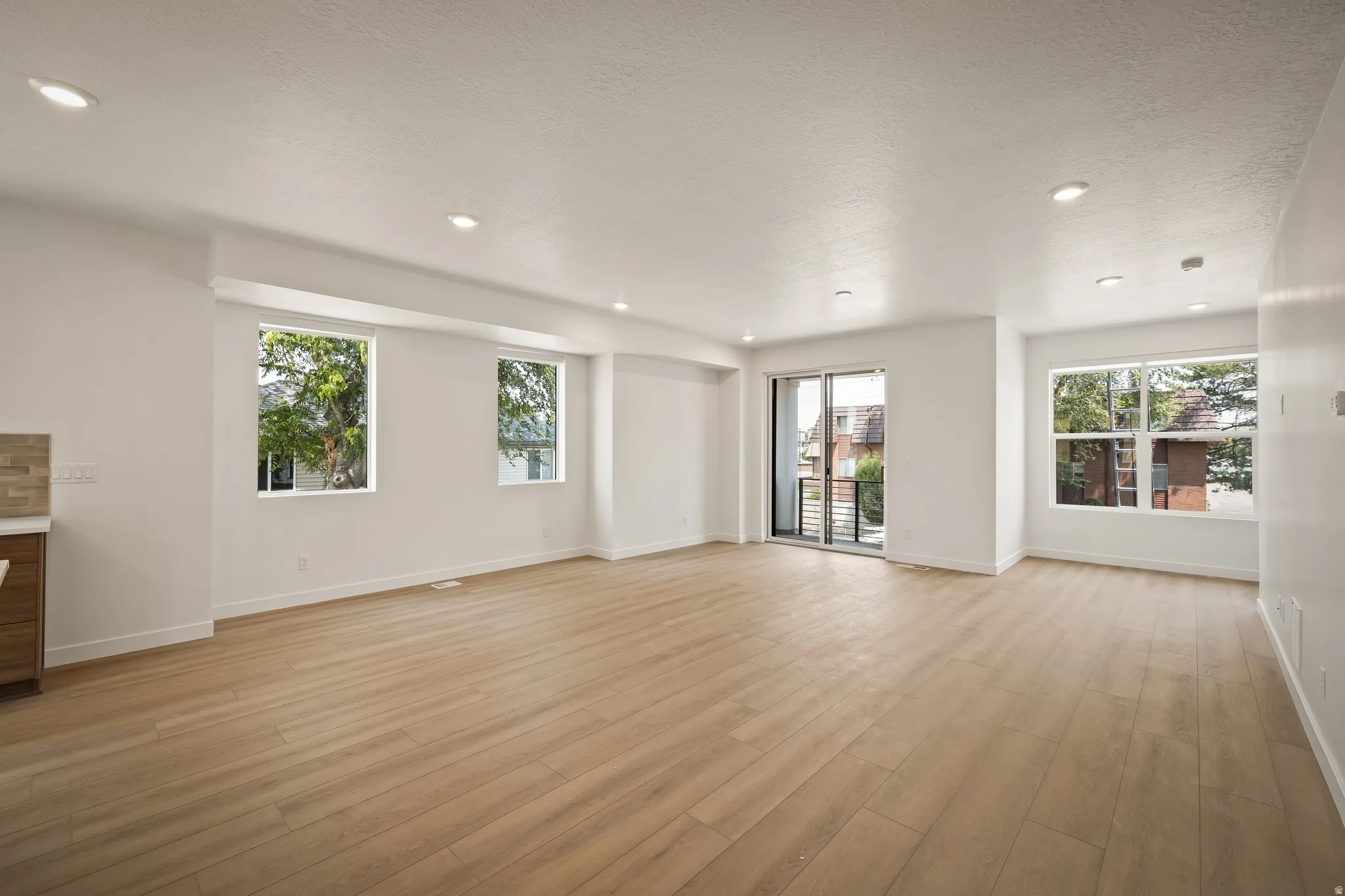 Expansive living area featuring light wood-finish flooring, recessed lighting, multiple windows, and a sliding glass door leading to a balcony