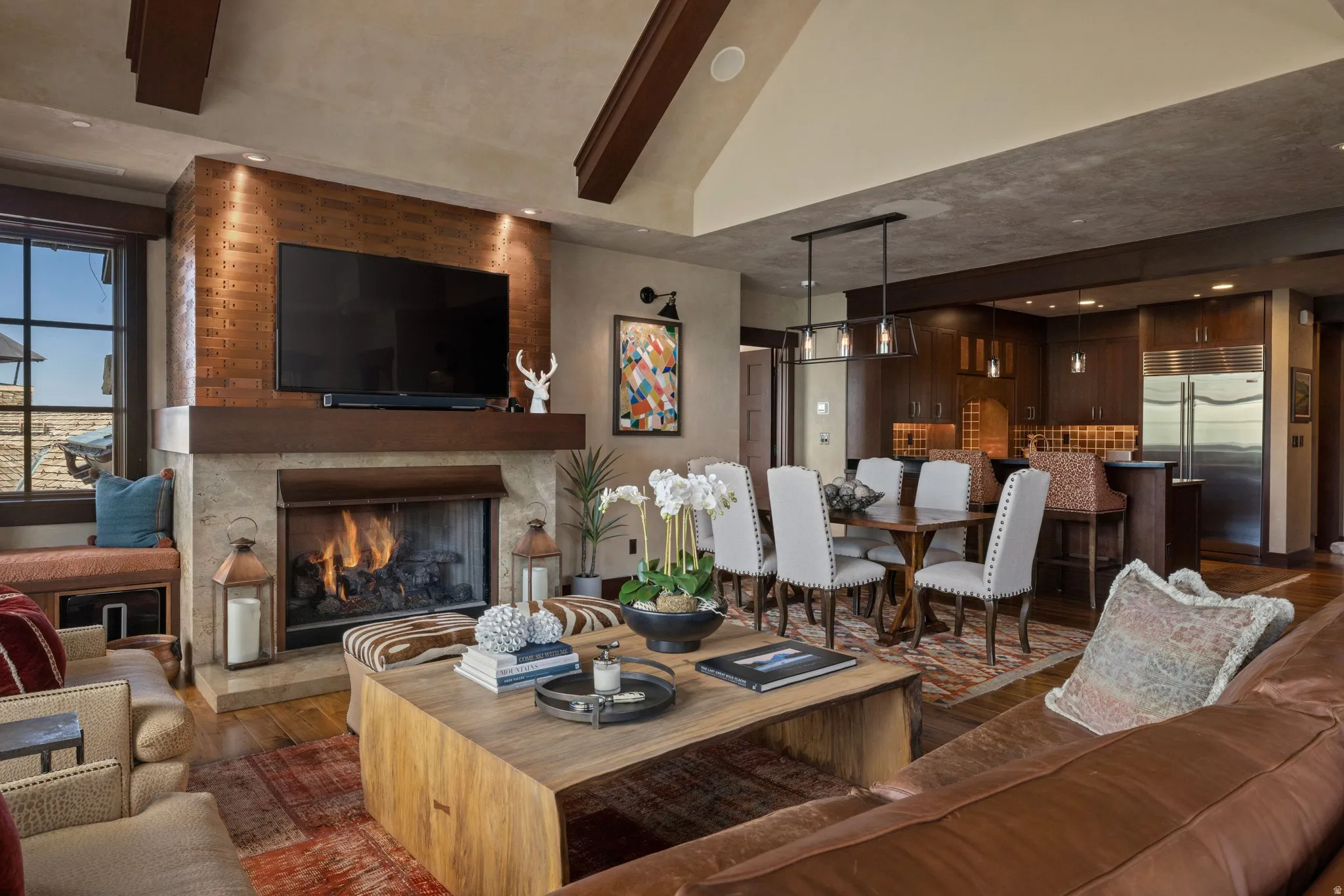 Exposed beam vaulted ceiling, stone fireplace with a wood mantel, wood-finish flooring, dining area with a linear chandelier, and a kitchen featuring built-in cabinetry and stainless steel appliances