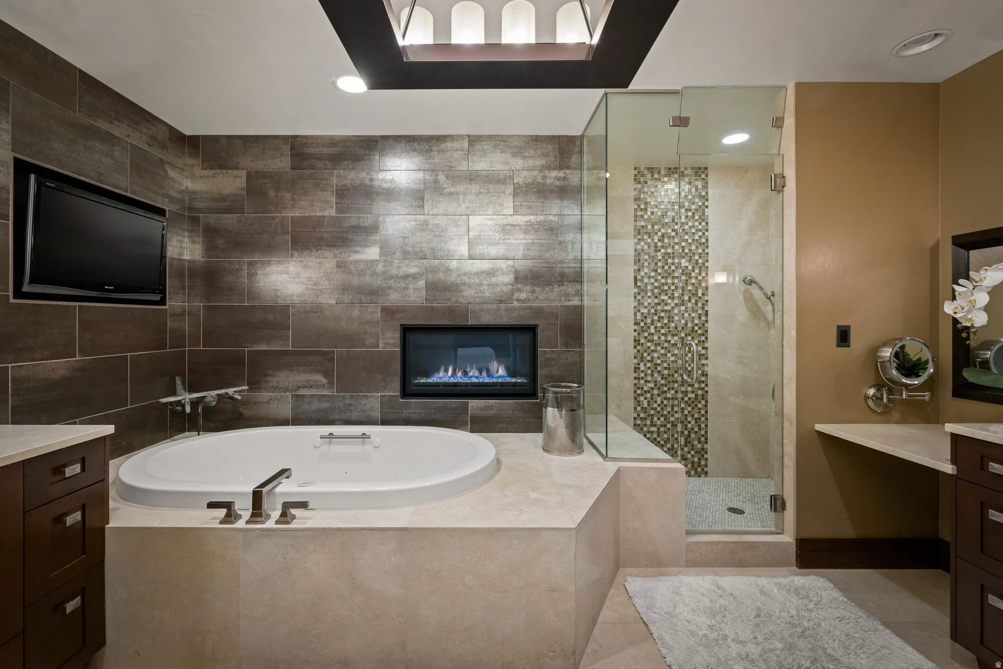 Spa-inspired bath featuring a built-in soaking tub, integrated fireplace, glass-enclosed shower with mosaic tile accent, and custom vanity cabinetry