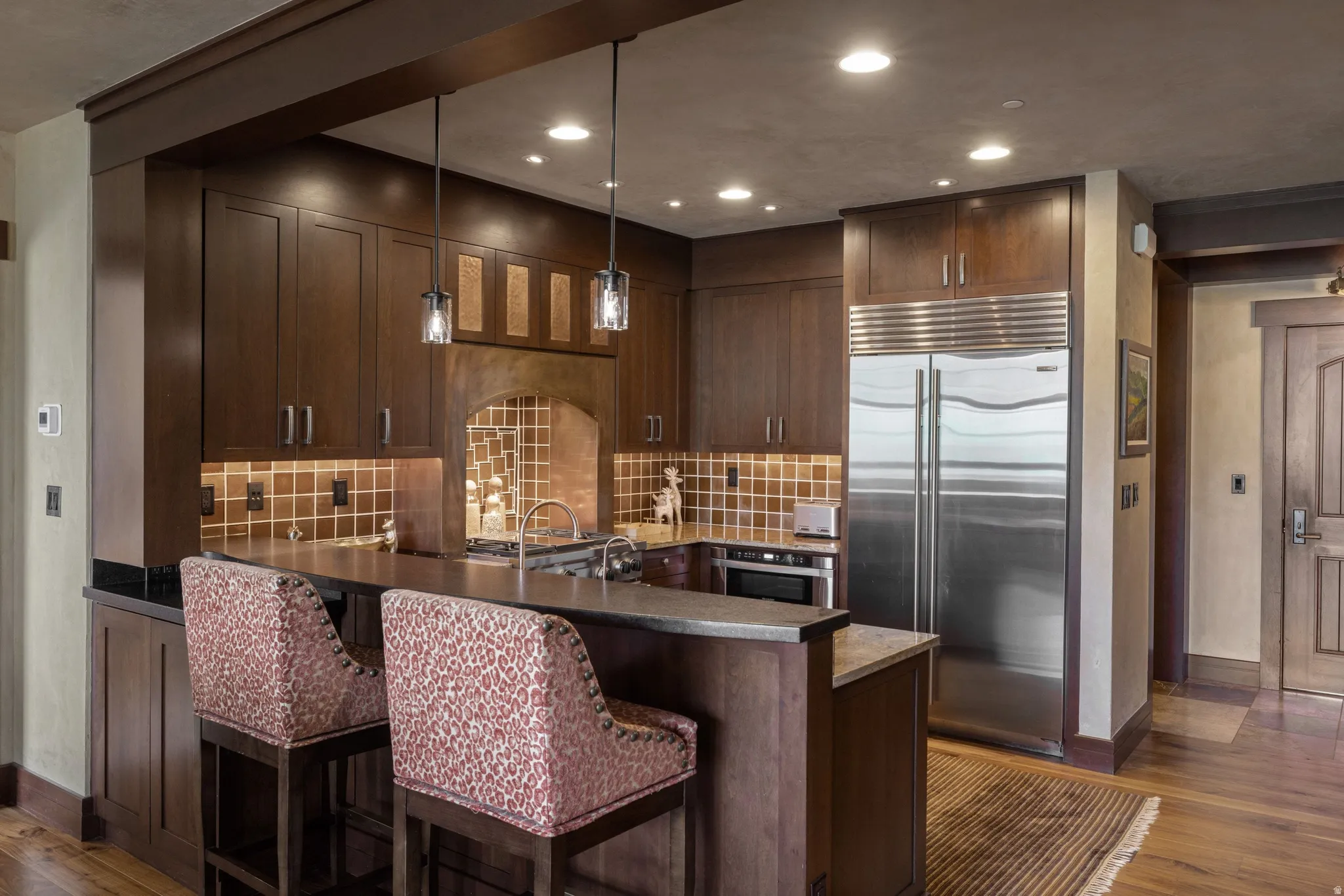 Rich wood cabinetry with integrated stainless steel appliances, a tiled backsplash, and recessed and pendant lighting