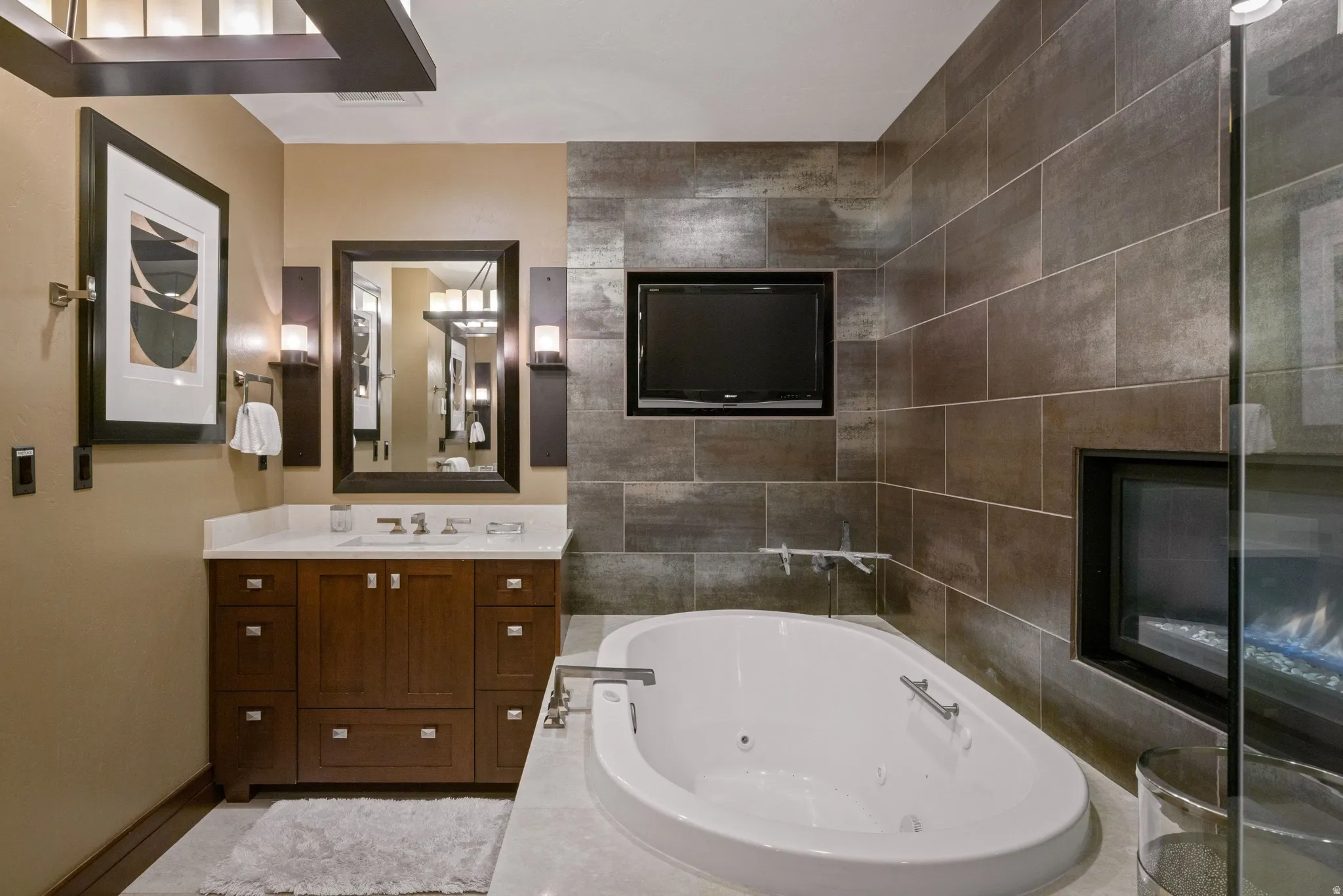 Deep soaking tub, built-in fireplace, wall-mounted television, wood vanity with integrated sink, and tiled accent wall