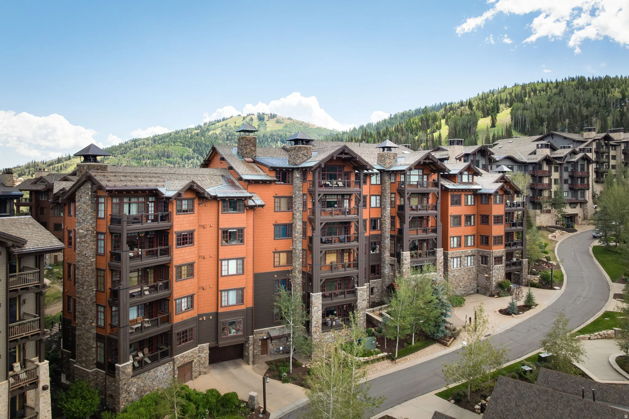 Multi-story residential building with stone and wood siding, multiple private balconies, a paved driveway, and surrounding mountainous terrain