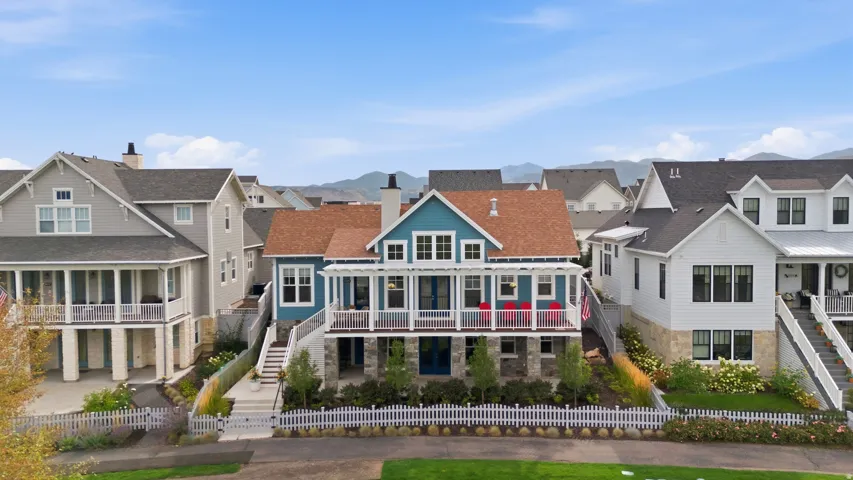 Coastal-inspired residence featuring a striking blue exterior, expansive covered porch with white railings, a brown shingle roof, and a stone foundation
