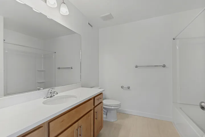 Light wood-finish flooring, a wood vanity with a white countertop, a large vanity mirror, and a white bathtub with a shower kit