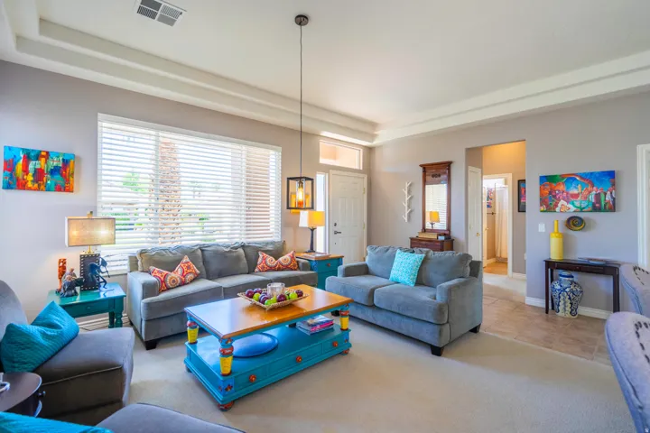Spacious formal living area featuring coffered ceiling details, large windows with plantation shutters, carpet, and a bright open layout with multiple seating zones and pendant lighting