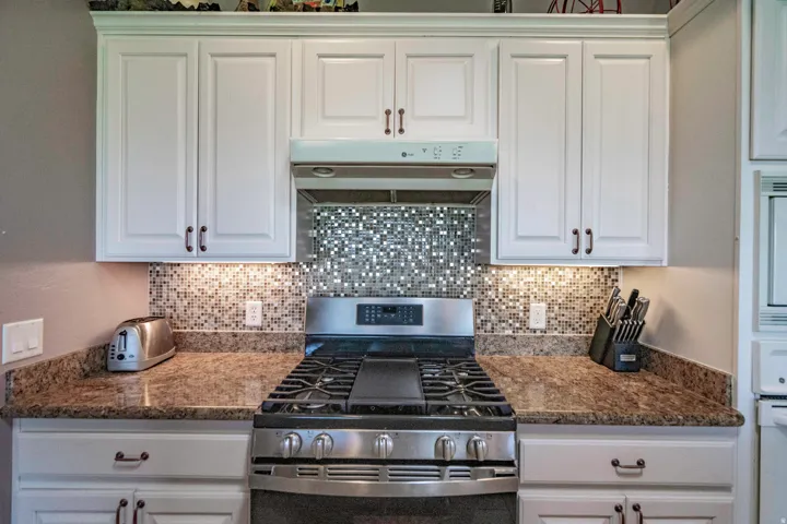 Chef's kitchen featuring custom white cabinetry, stainless steel gas range with six burners, mosaic tile backsplash, and granite countertops with ample workspace and storage throughout