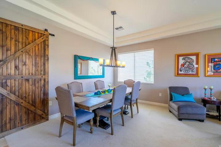 Spacious open-concept formal living and dining area featuring a dining table with upholstered chairs, pendant lighting with brass accents, large windows with plantation shutters, and neutral wall finishes. Sliding barn door.