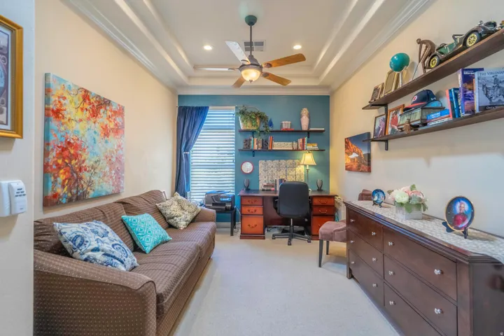 Versatile bedroom retreat featuring coffered ceiling with recessed lighting and ceiling fan, built-in desk workspace with wooden cabinetry and ample storage, floating shelving with accent lighting, and coordinating dresser with multiple drawers