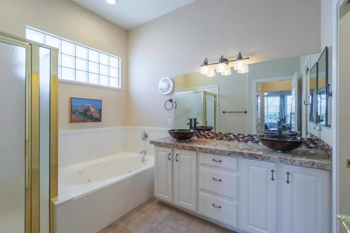 Dual vessel sinks set on a stone-finish countertop, full vanity mirror, built-in bathtub, and a glass-block window