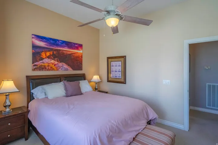 Spacious 2nd bedroom with wood-finish furnishings, dual bedside table lamps with ceramic bases, layered bedding in soft purple tones, ceiling fan with integrated lighting, and large scenic artwork above the headboard