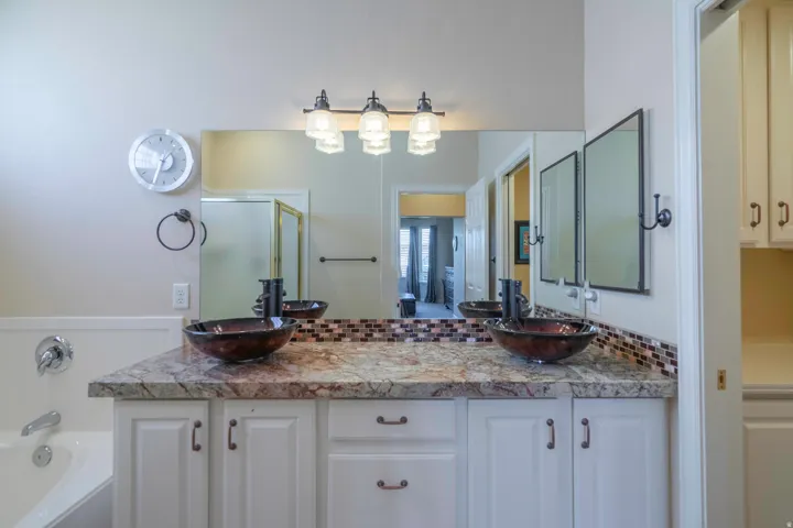 Dual vessel sinks, stone-finish countertop, white cabinetry with bronze hardware, mosaic tile backsplash, and a built-in bathtub