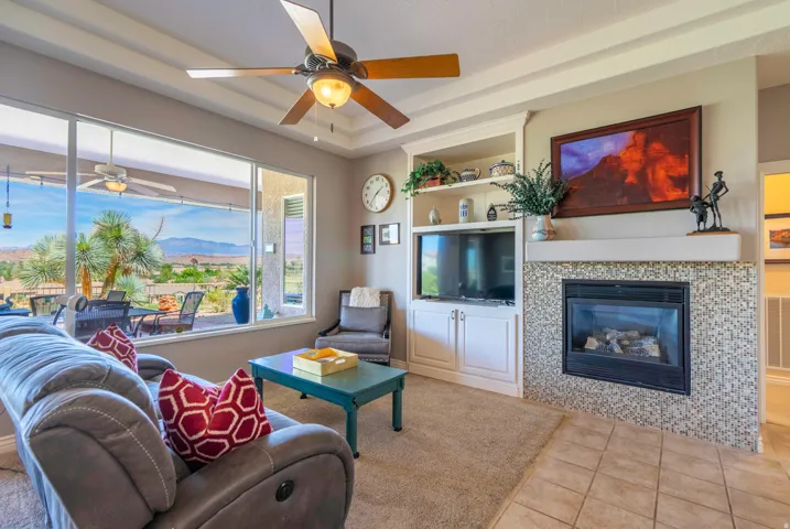 Bright, open living space featuring expansive windows with scenic outlooks, carpet covered tile flooring, a wood toned ceiling fan with integrated lighting, built-in cabinetry with display shelving, a gas fireplace with decorative tile surround, and a wal