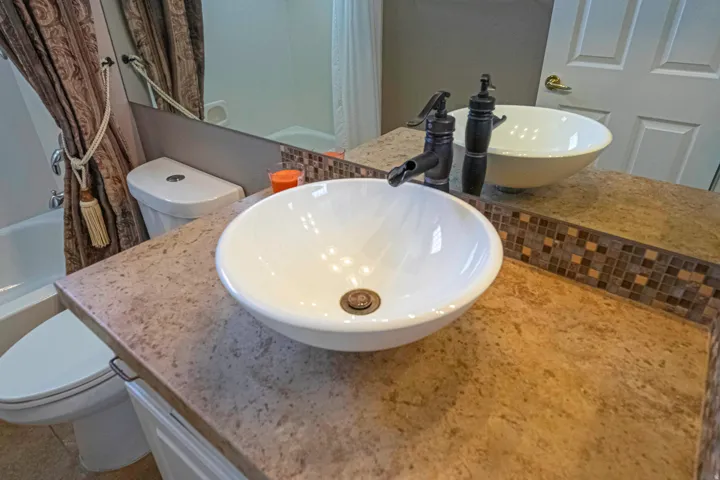 Dual vessel sink vanity featuring round white ceramic basins, warm-toned stone countertops with decorative tile backsplash, and matte black faucet fixtures