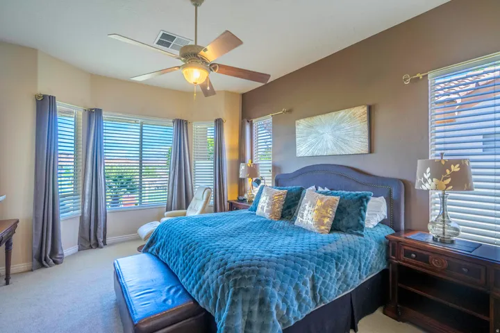 Master bedroom featuring a ceiling fan with integrated lighting, expansive windows with plantation shutters providing natural light and privacy control, neutral wall finishes, and wood-finish nightstands with attached lighting fixtures