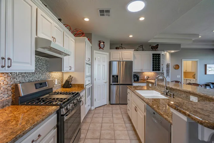 Chef's kitchen featuring stainless steel appliances, dual gas cooktop with professional-grade range, custom white cabinetry with hardware accents, granite countertops with waterfall island edge, mosaic tile backsplash, recessed lighting, and tile flooring